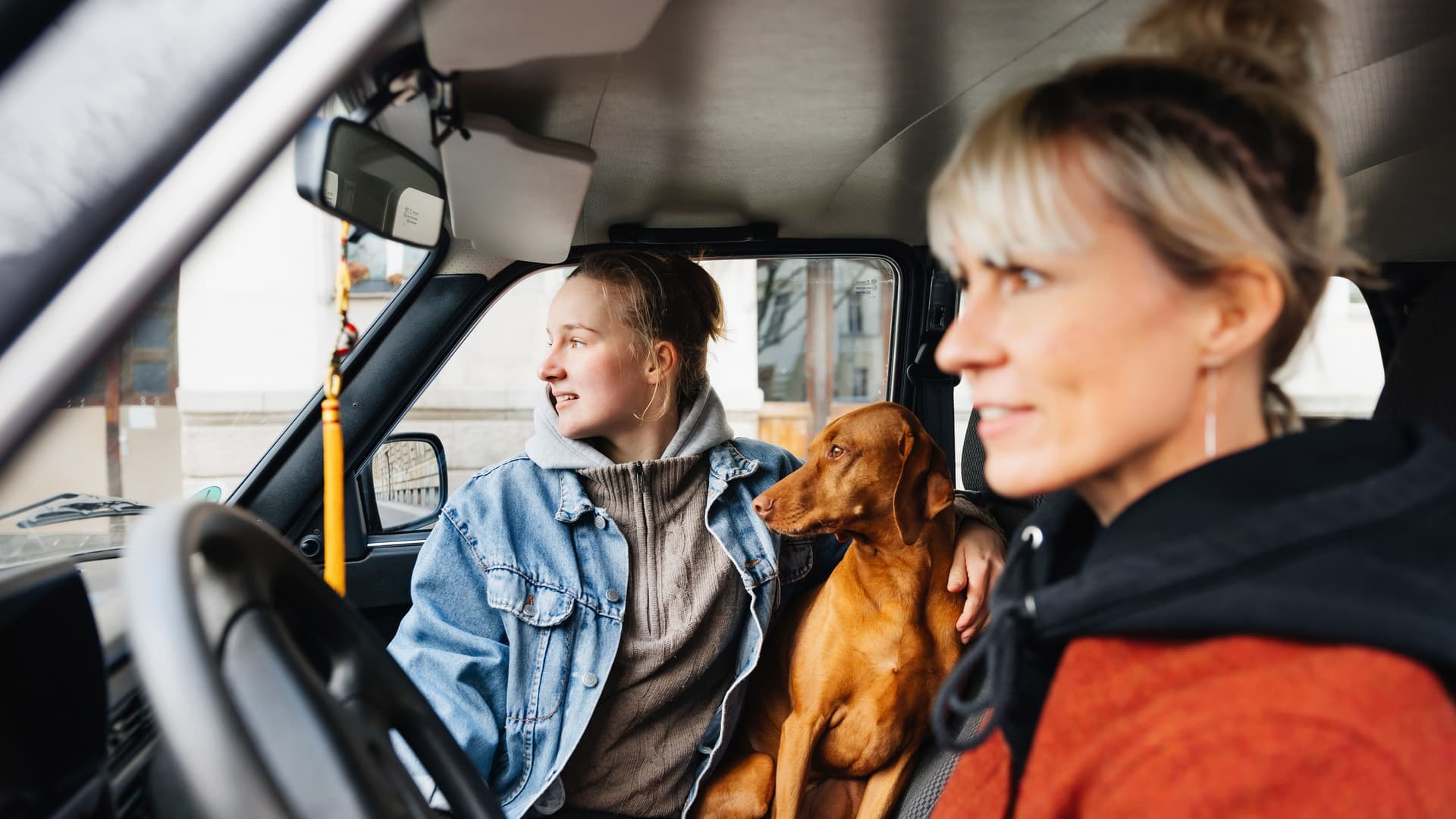 Eine Frau sitzt am Steuer eines Vans während ein Mädchen im Teenageralter und ihr Hund neben ihr sitzen.